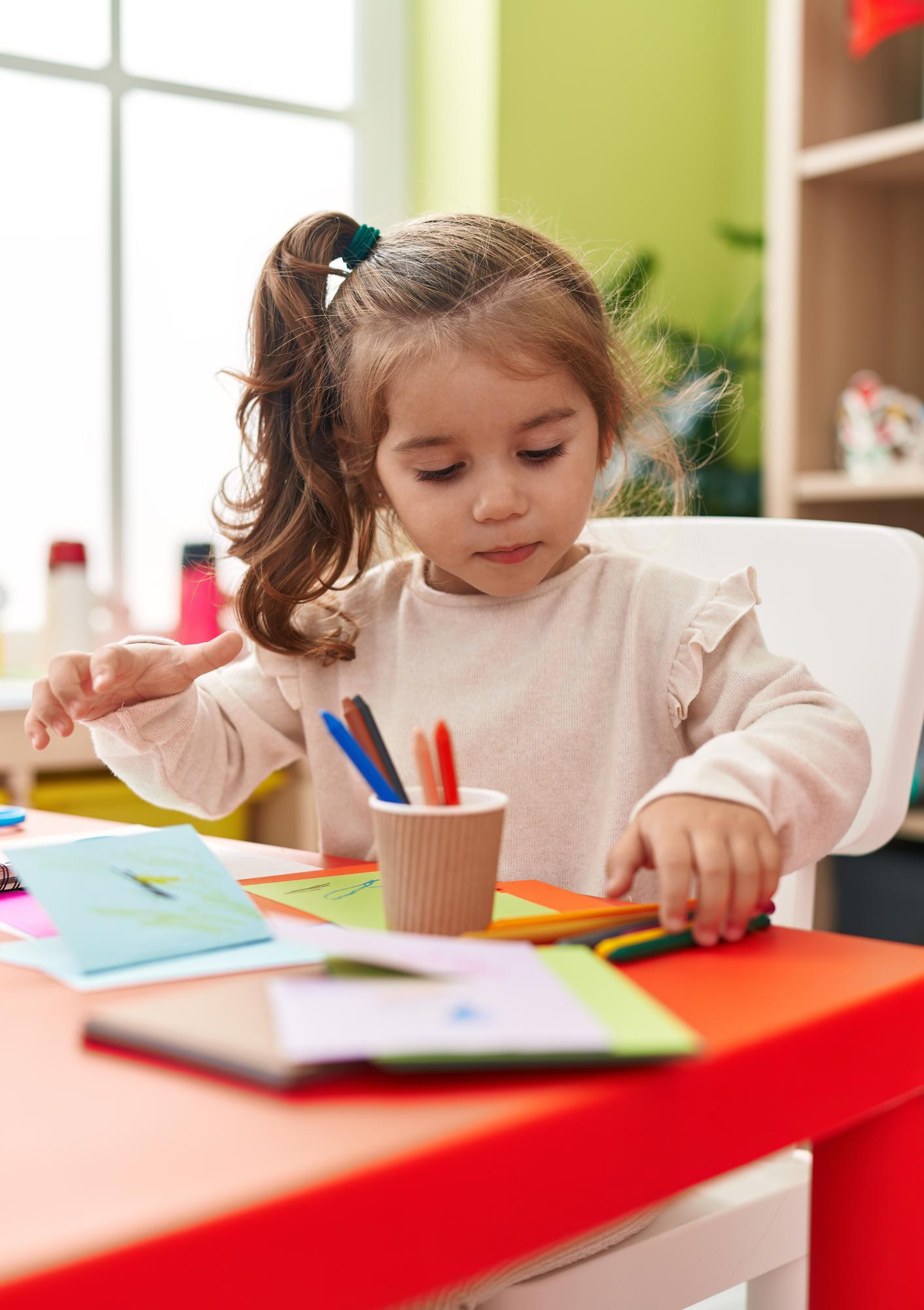 Toddler girl sitting it classroom setting on a red desk, colors on a card using colored pencils.