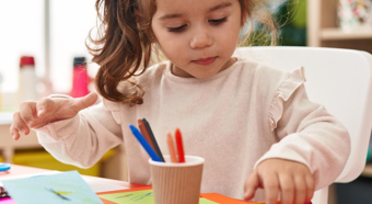 Toddler girl sitting it classroom setting on a red desk, colors on a card using colored pencils.