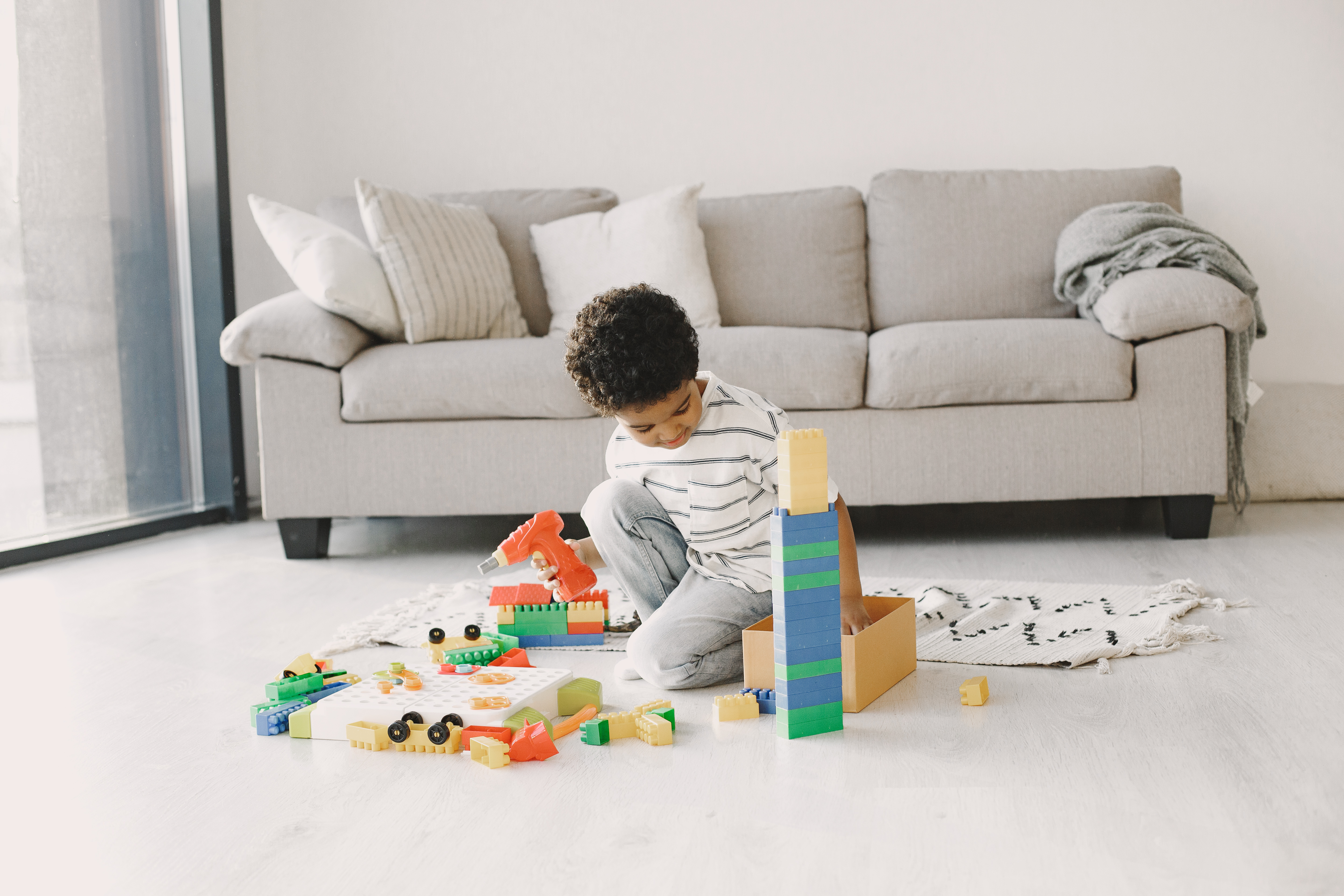 Toddler boy sits in middle of a living room, playing with various toys and blocks.