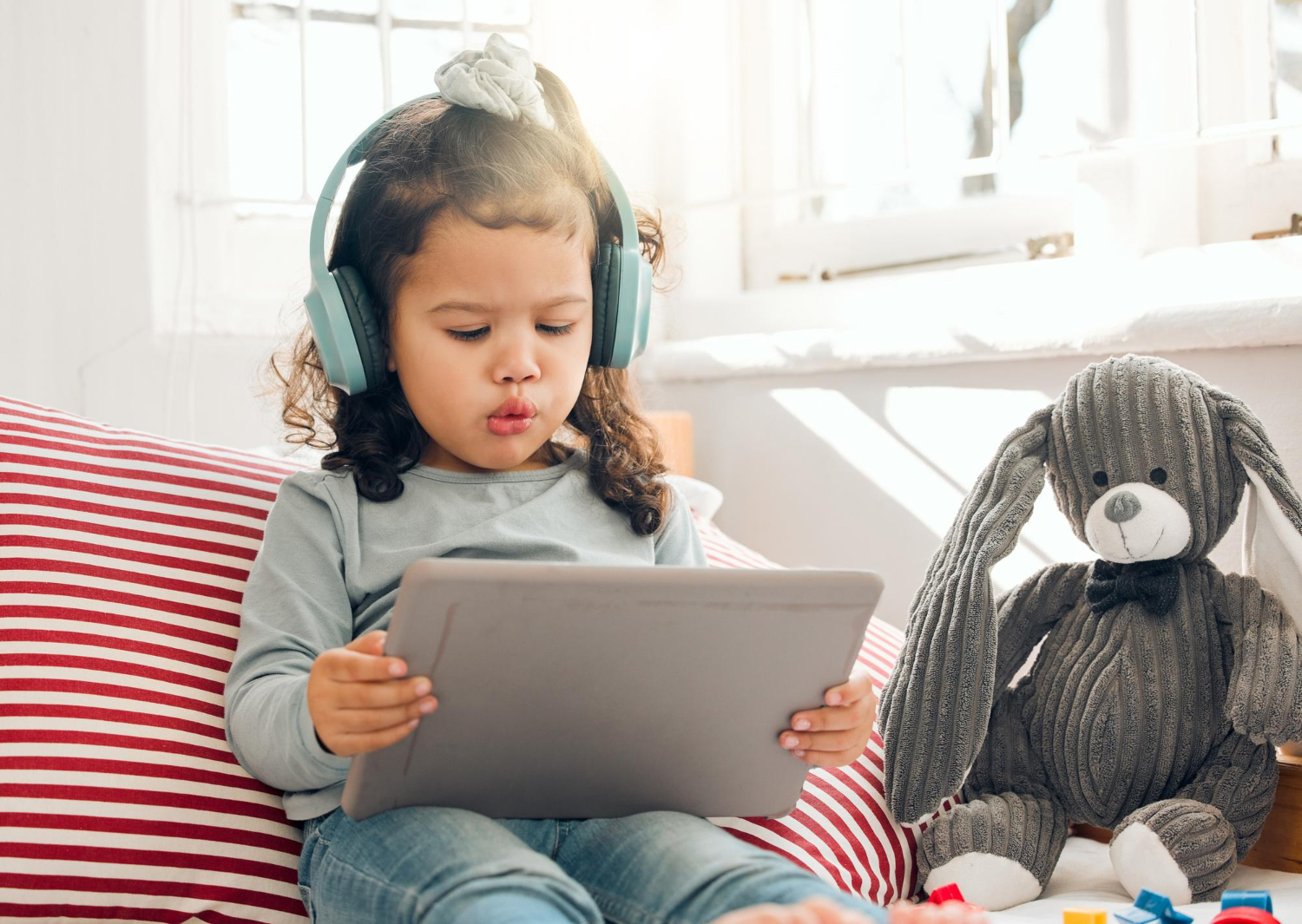 Toddler girl sitting on a home couch setting looks at iPad with big headphones on her head. 