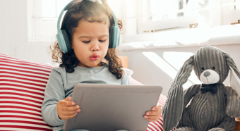Toddler girl sitting on a home couch setting looks at iPad with big headphones on her head. 