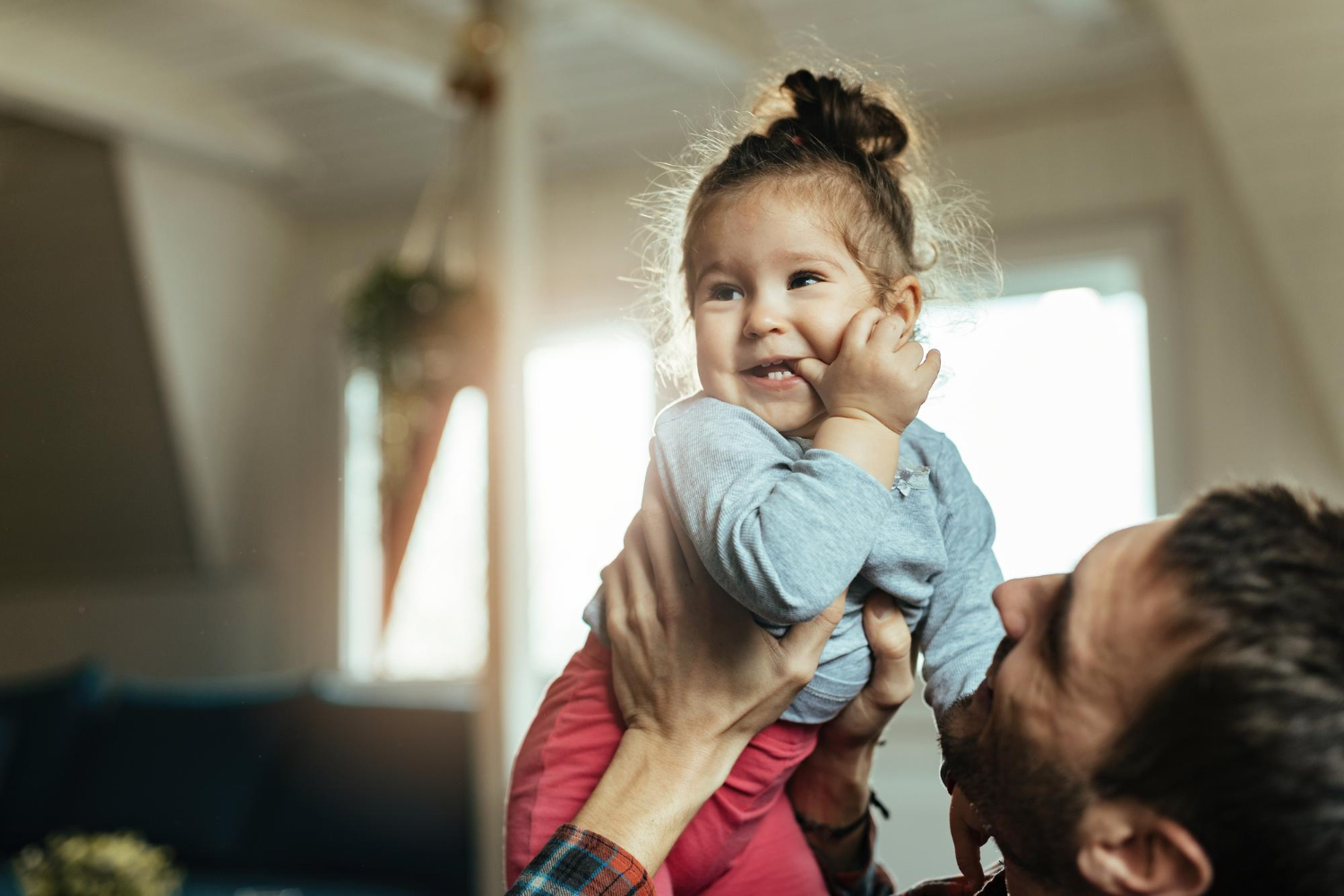 Adult male holds up little girl in air. Both smiling, in an indoor house setting.