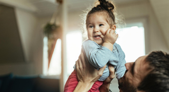 Adult male holds up little girl in air. Both smiling, in an indoor house setting.