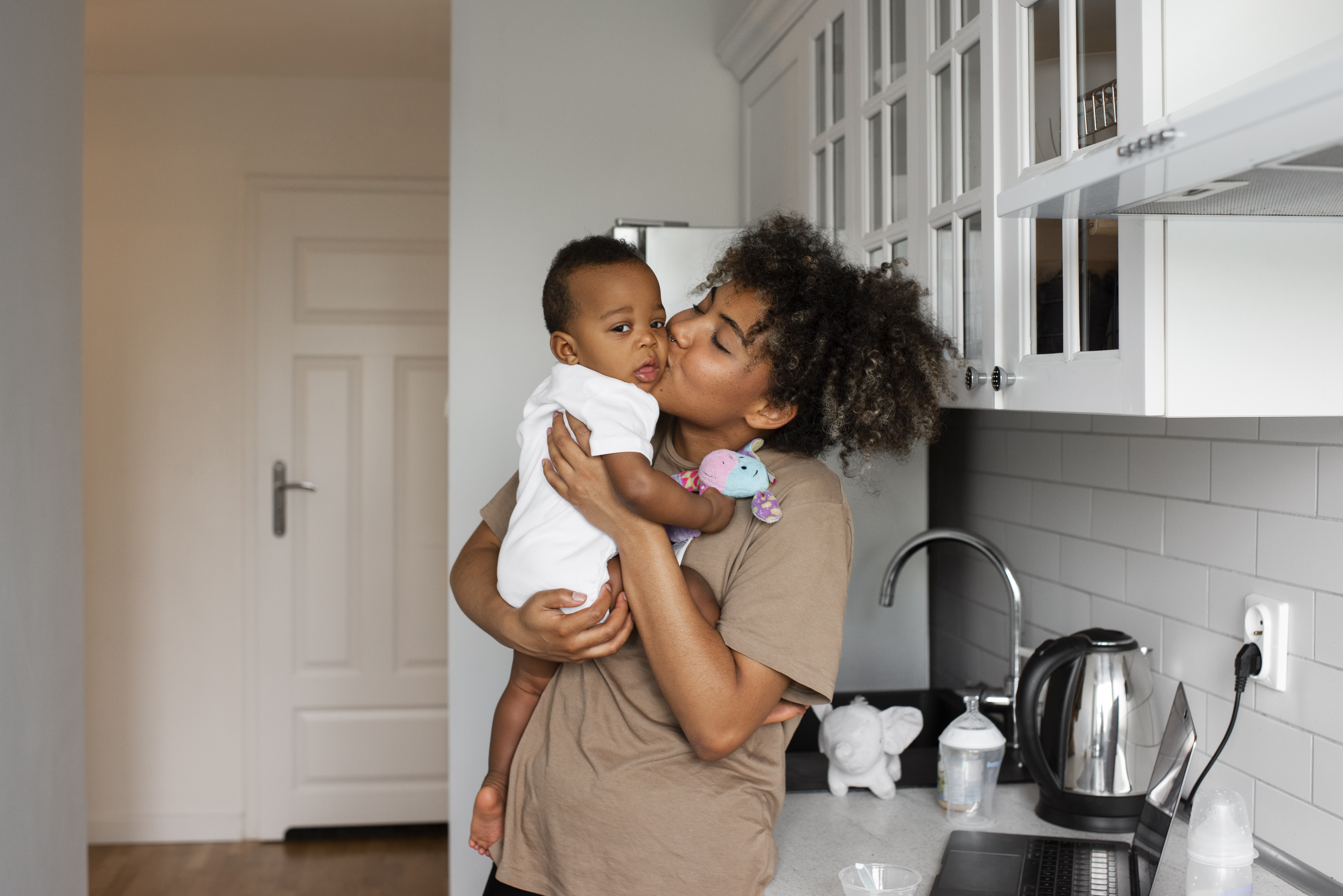 Young women holds toddler and gives him a kiss on the cheek in a home kitchen setting.