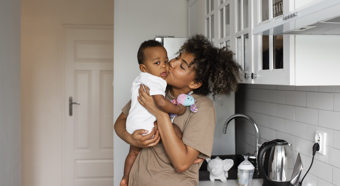 Young women holds toddler and gives him a kiss on the cheek in a home kitchen setting.