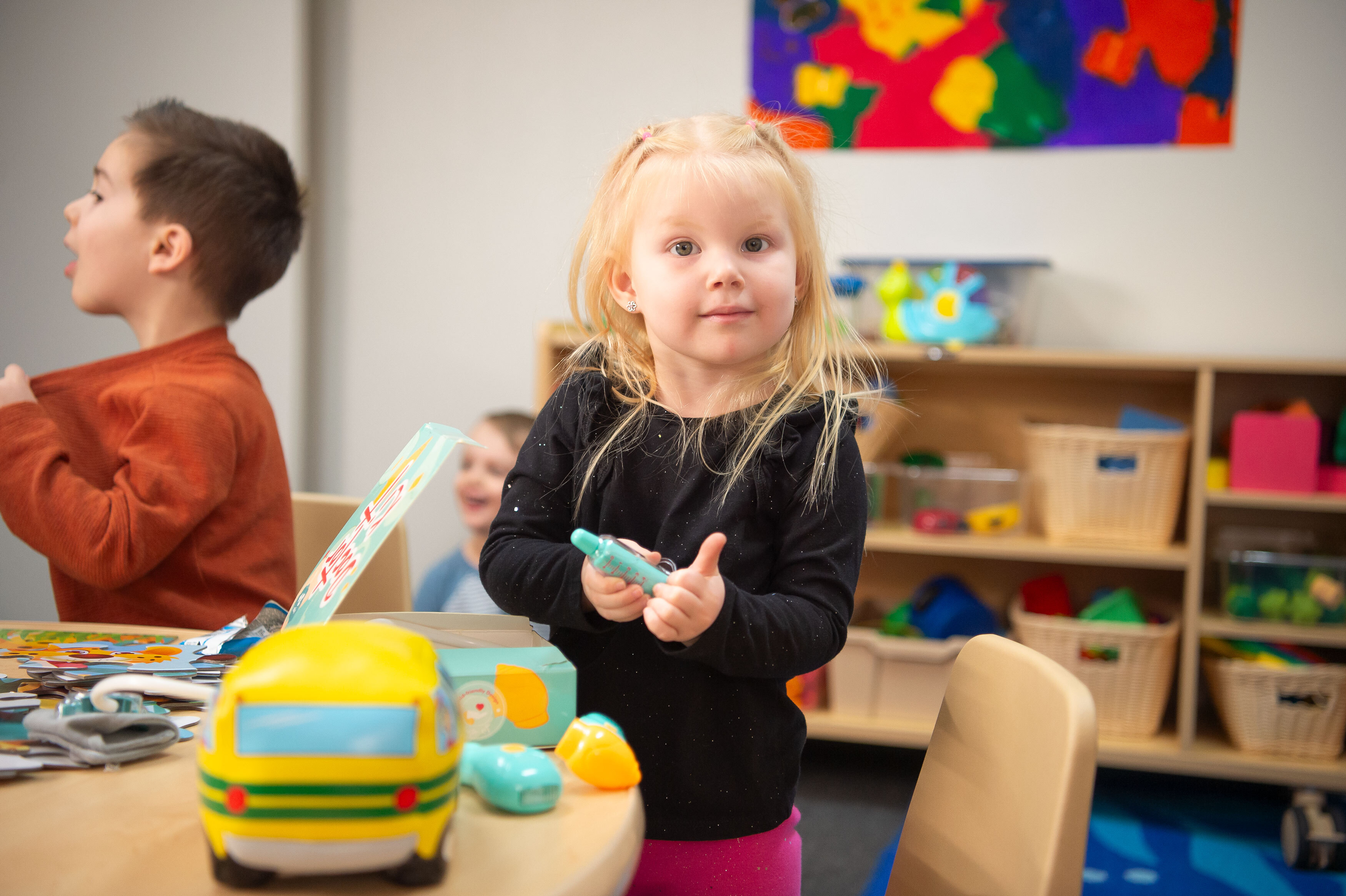 Young girl and boy with light skin and hair in a classroom playing with toys at a table.