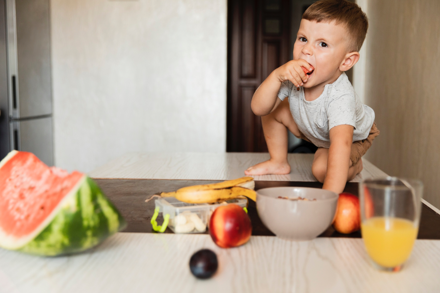 Toddler in a home kitchen setting eats a fruit. There is also a watermelon, banana, apple, orange juice and other fruits on the table. 
