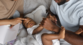 Two adult figures sit in bed with baby, both smiling and comforting the baby in a comfy setting. 