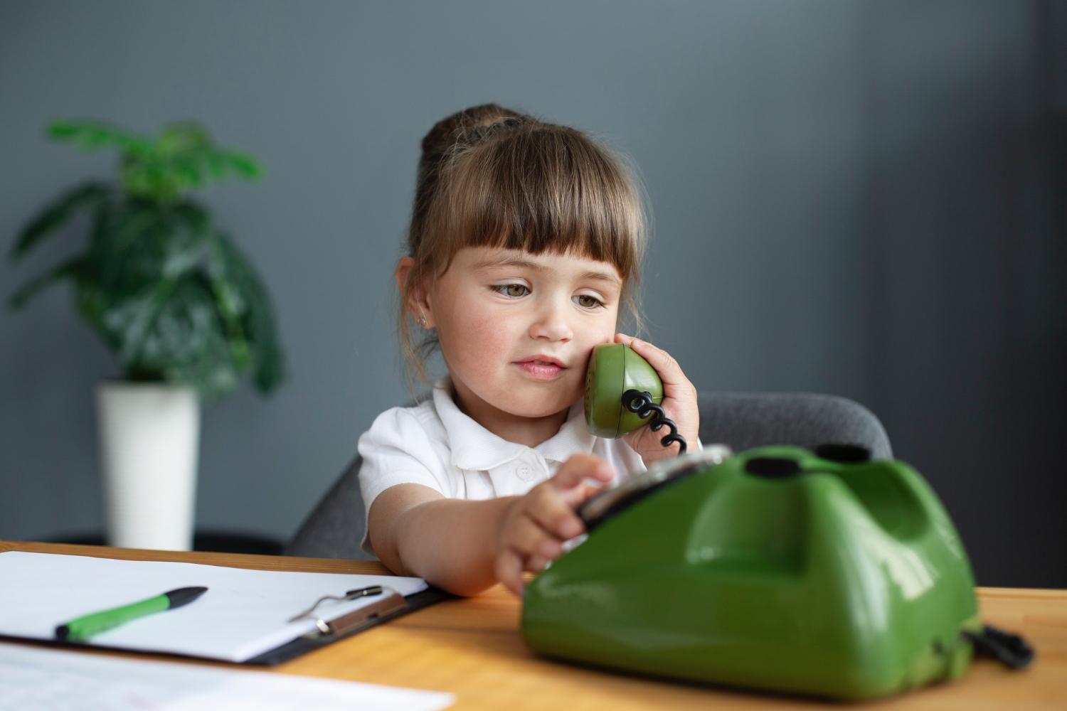 Young toddler girl dressed in nice clothes pretending to talk on a green phone, with paperwork on desk next to her, plant in background. 