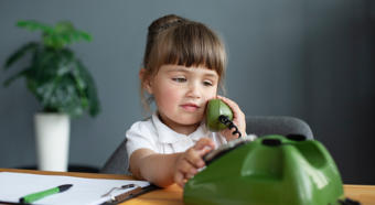Young toddler girl dressed in nice clothes pretending to talk on a green phone, with paperwork on desk next to her, plant in background. 