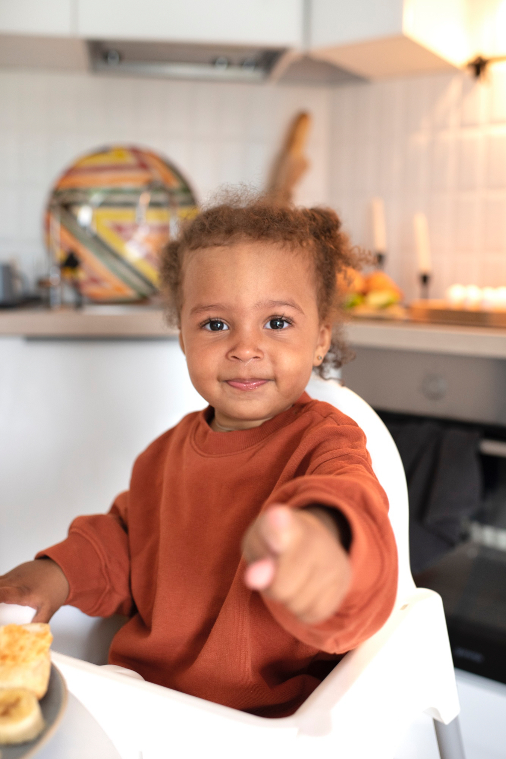 Happy little girl smiles and points right at the camera. In an indoor kitchen setting, sitting in her highchair. 