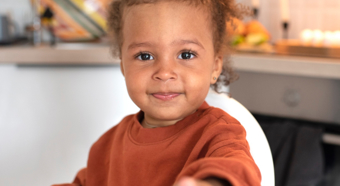 Happy little girl smiles and points right at the camera. In an indoor kitchen setting, sitting in her highchair. 