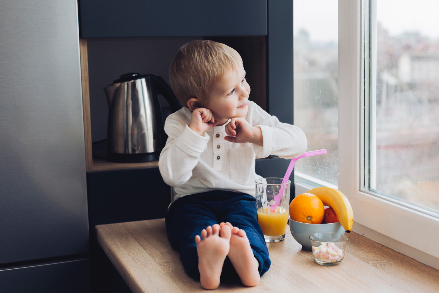 Little boy sitting in window sill, smiling and looking outside. Orange juice with a pink Straw and bowl of fruit are next to him. 