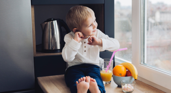 Little boy sitting in window sill, smiling and looking outside. Orange juice with a pink Straw and bowl of fruit are next to him. 
