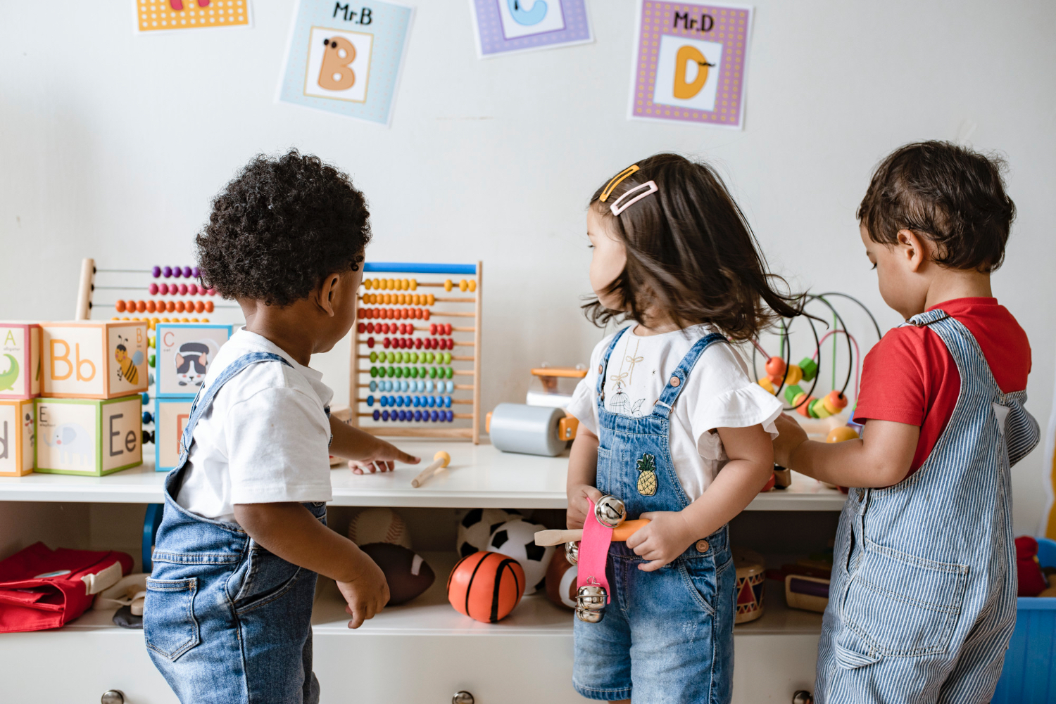 Three toddlers all wearing jean overalls play with toys together in a classroom setting.