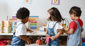 Three toddlers all wearing jean overalls play with toys together in a classroom setting.