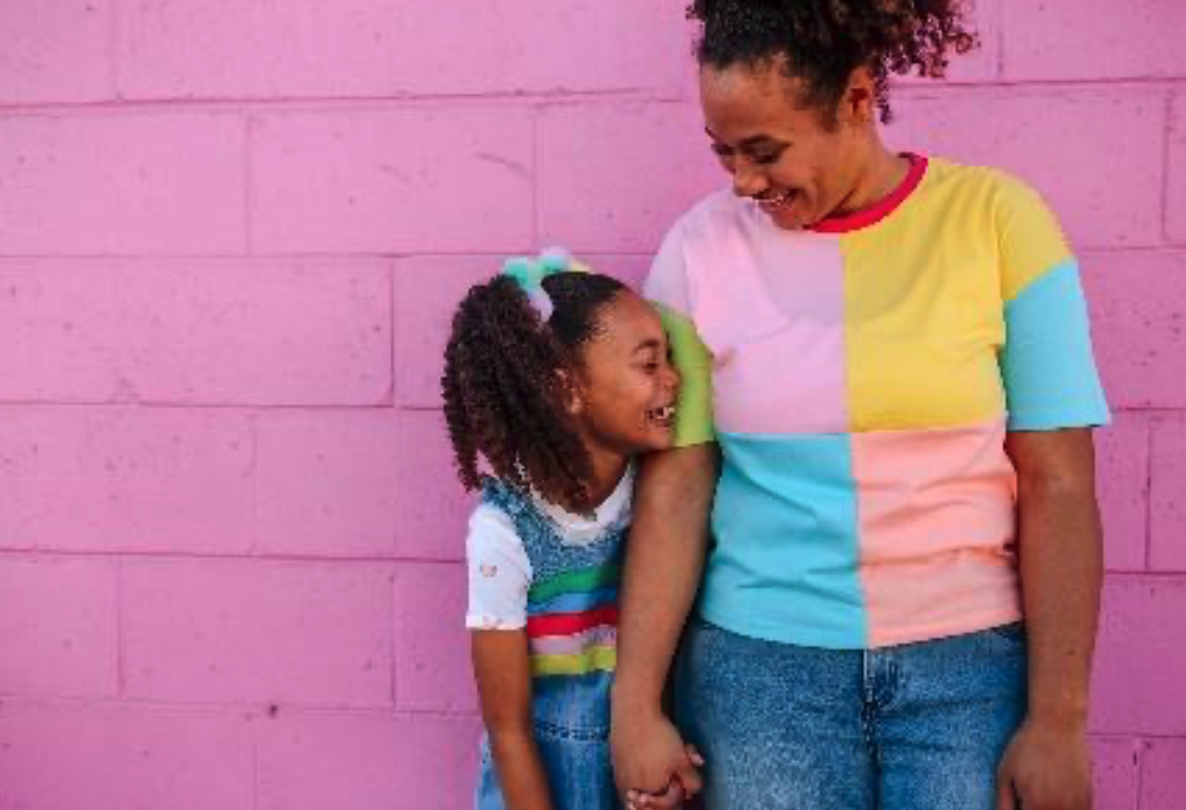 Adult female holding hands with young girl laughing and smiling at each other, standing in front of a pink wall, both wearing colorful clothing. 