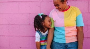 Adult female holding hands with young girl laughing and smiling at each other, standing in front of a pink wall, both wearing colorful clothing. 