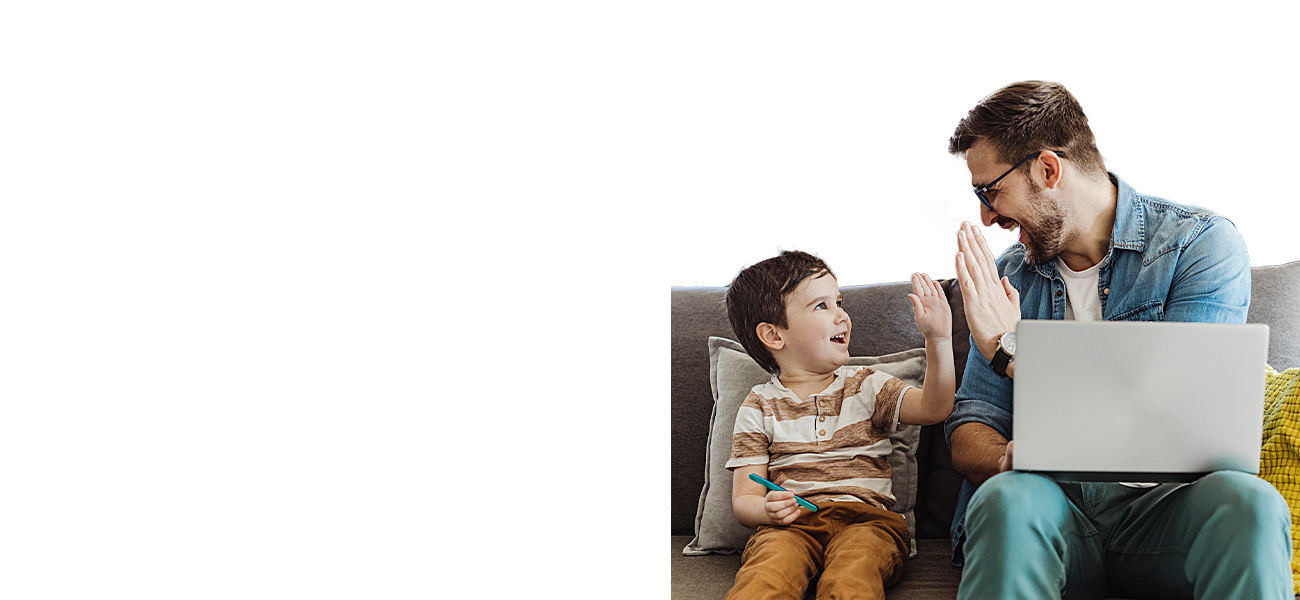 An adult male with light skin and short-styled brown hair is high-fiving a young boy with light skin and short brown hair while they both sit on a grey couch. The man is holding a computer on his lap.