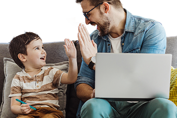 An adult male with light skin and short-styled brown hair is high-fiving a young boy with light skin and short brown hair while they both sit on a grey couch. The man is holding a computer on his lap.