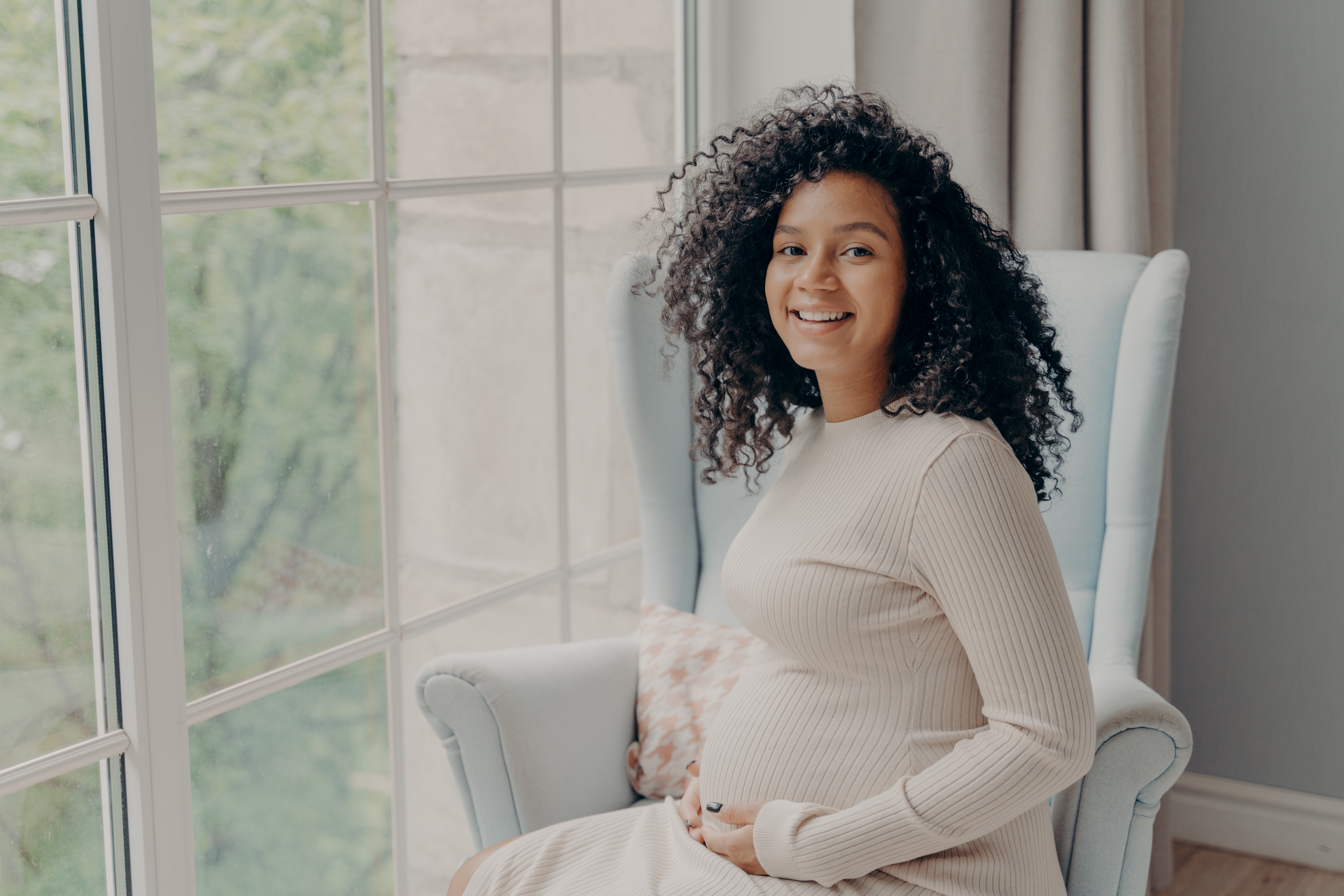 A pregnant woman with light brown skin and curly long hair poses on a chair to show her belly.