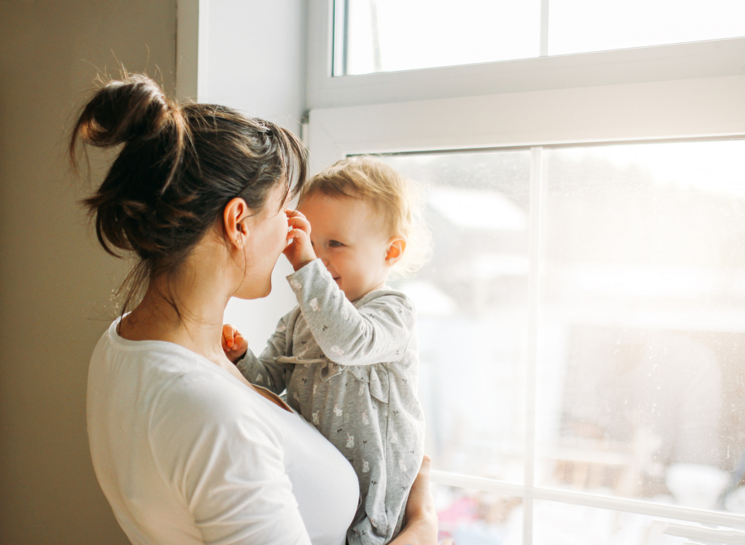 Adult female holding toddler in bright indoor setting. Toddler is smiling and playing with the adults nose.