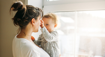 Adult female holding toddler in bright indoor setting. Toddler is smiling and playing with the adults nose.
