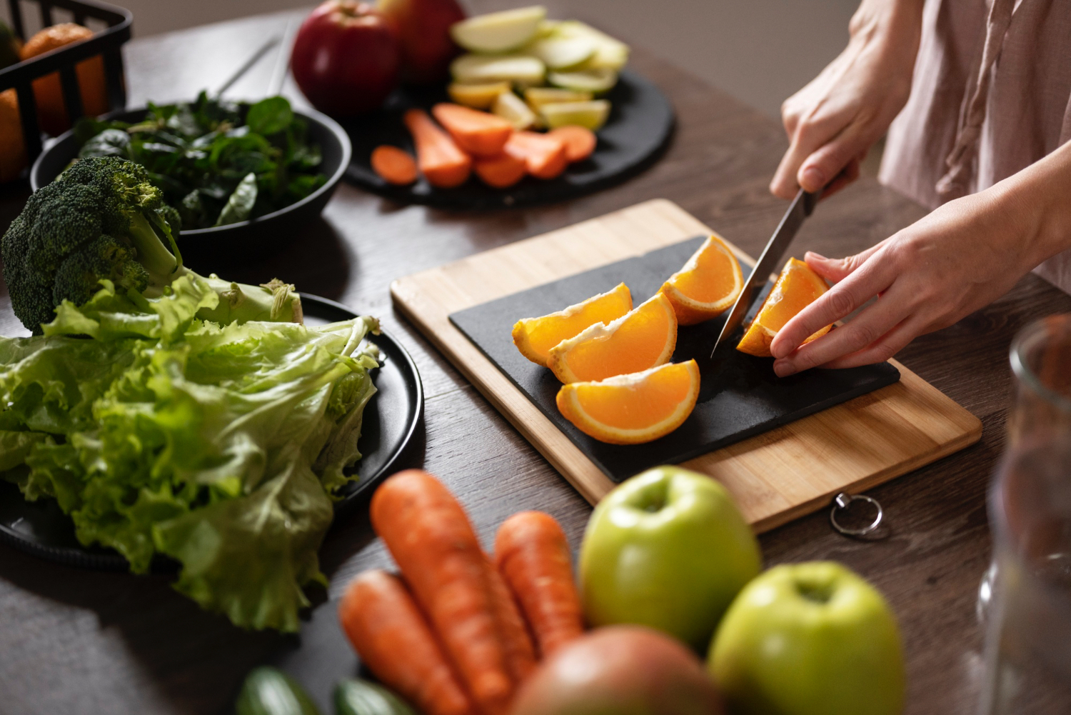 Kitchen setting where person is cutting various fruits and vegetables. Lettuce, carrots, oranges, apples broccoli 