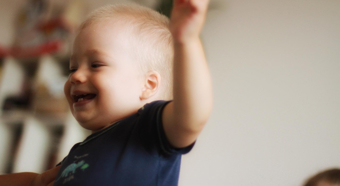 Blonde baby with short hair looks to left while smiling and holding his hand up in the air. 