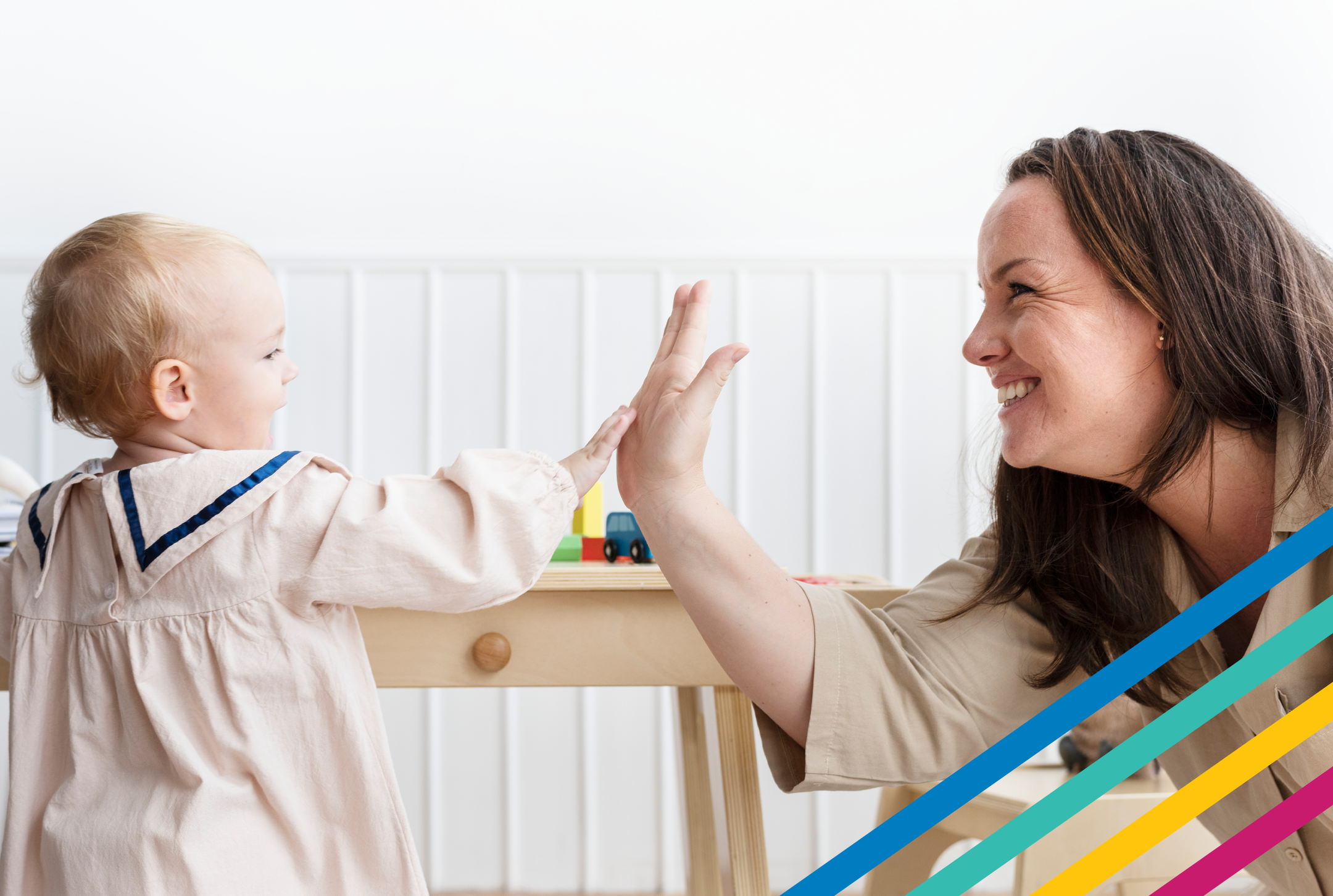 Adult female high-fives a toddler wearing a light pink dress in a bright home setting with toddler toys in background.