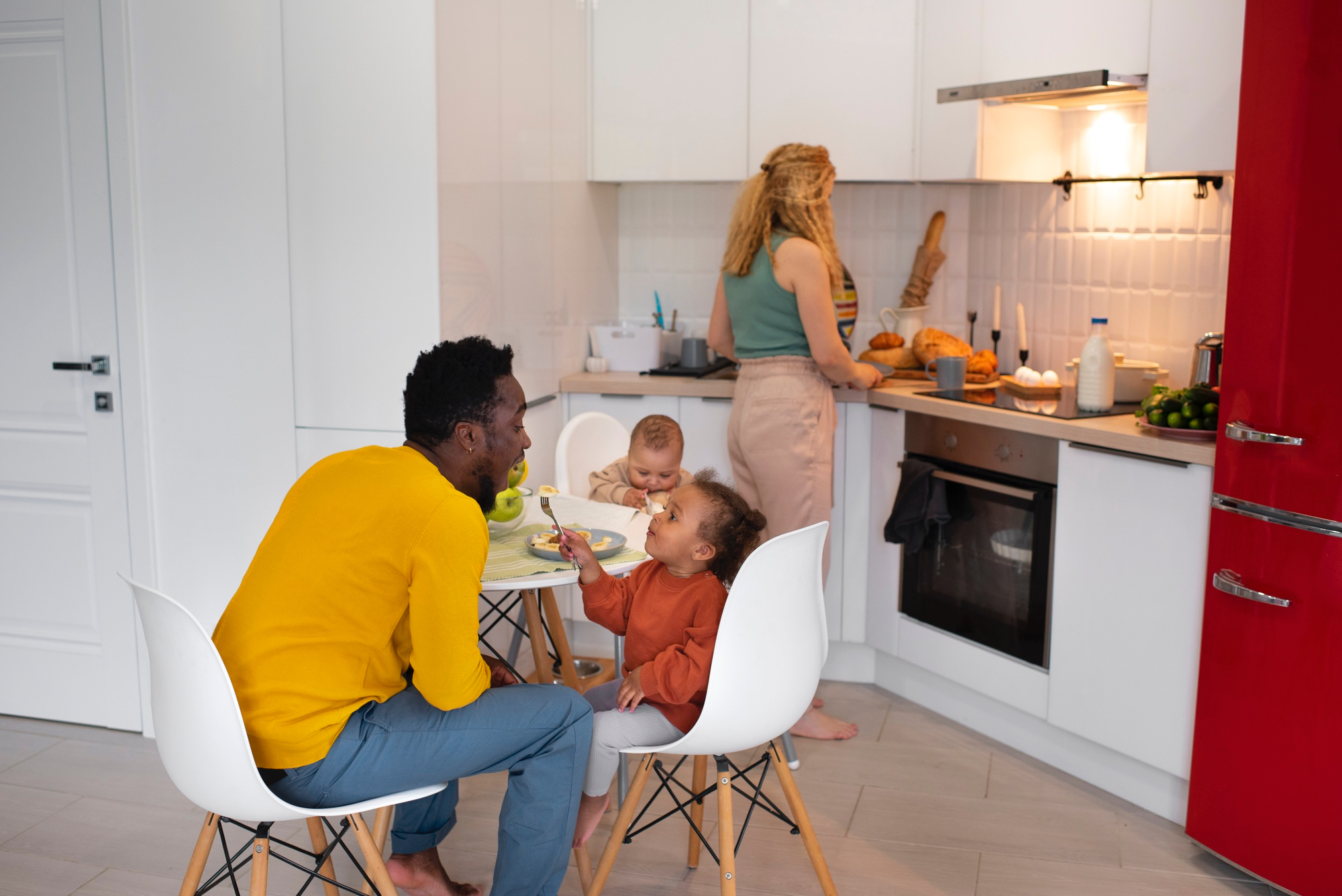 Adult female and male in a kitchen setting, with two toddlers, one sitting at the table with a fork in hand and the other in a high chair. Female cooking. 