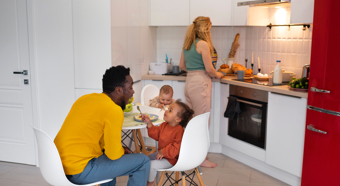 Adult female and male in a kitchen setting, with two toddlers, one sitting at the table with a fork in hand and the other in a high chair. Female cooking. 