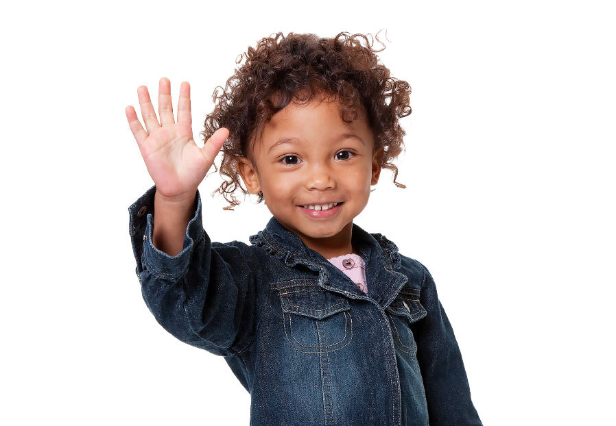A young girl with brown skin and curly brown hair is wearing a jean jacket and smiling at the camera while holding her hand up and waving.