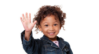 A young girl with brown skin and curly brown hair is wearing a jean jacket and smiling at the camera while holding her hand up and waving.