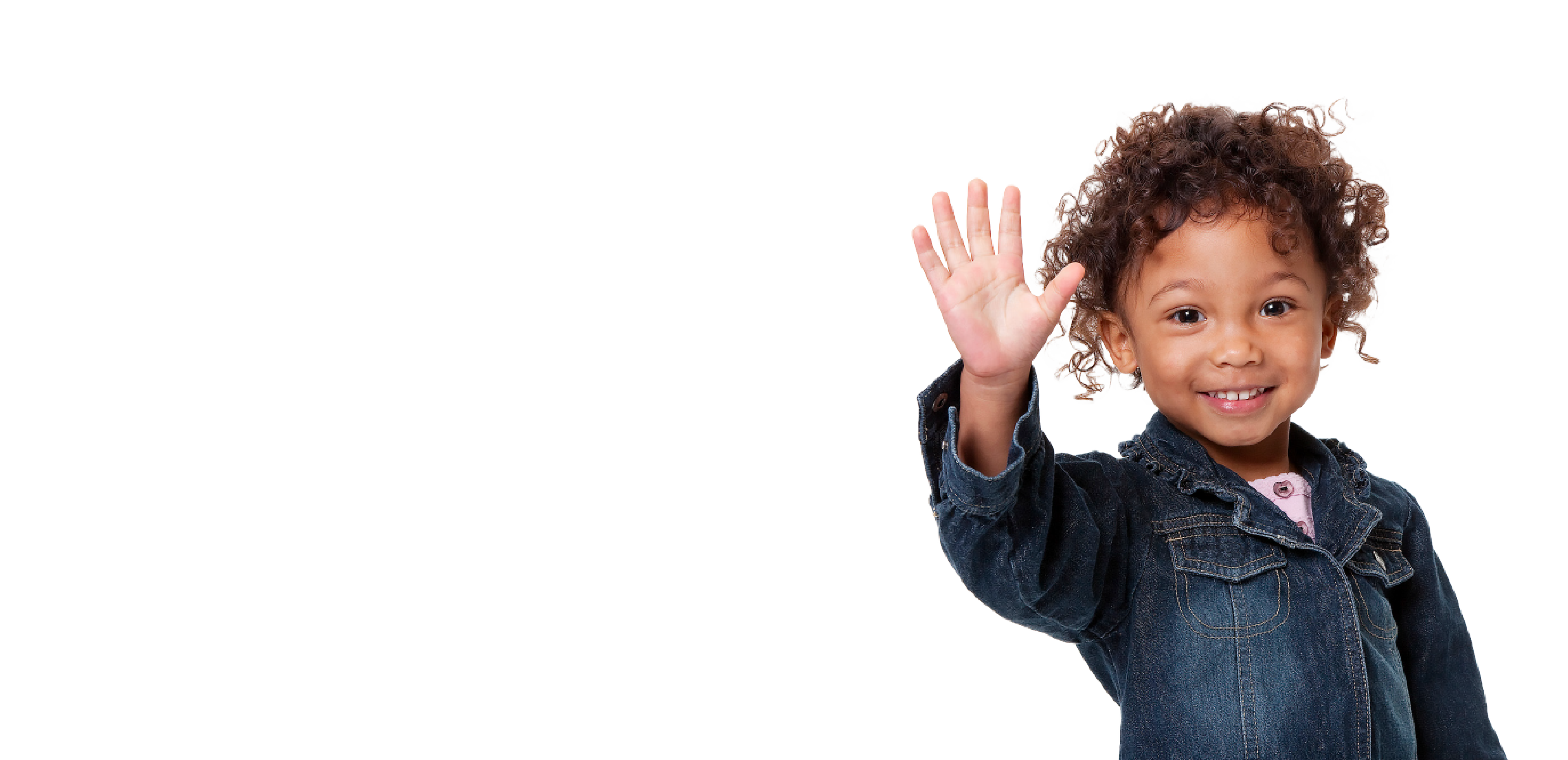 A young girl with brown skin and curly brown hair is wearing a jean jacket and smiling at the camera while holding her hand up and waving.