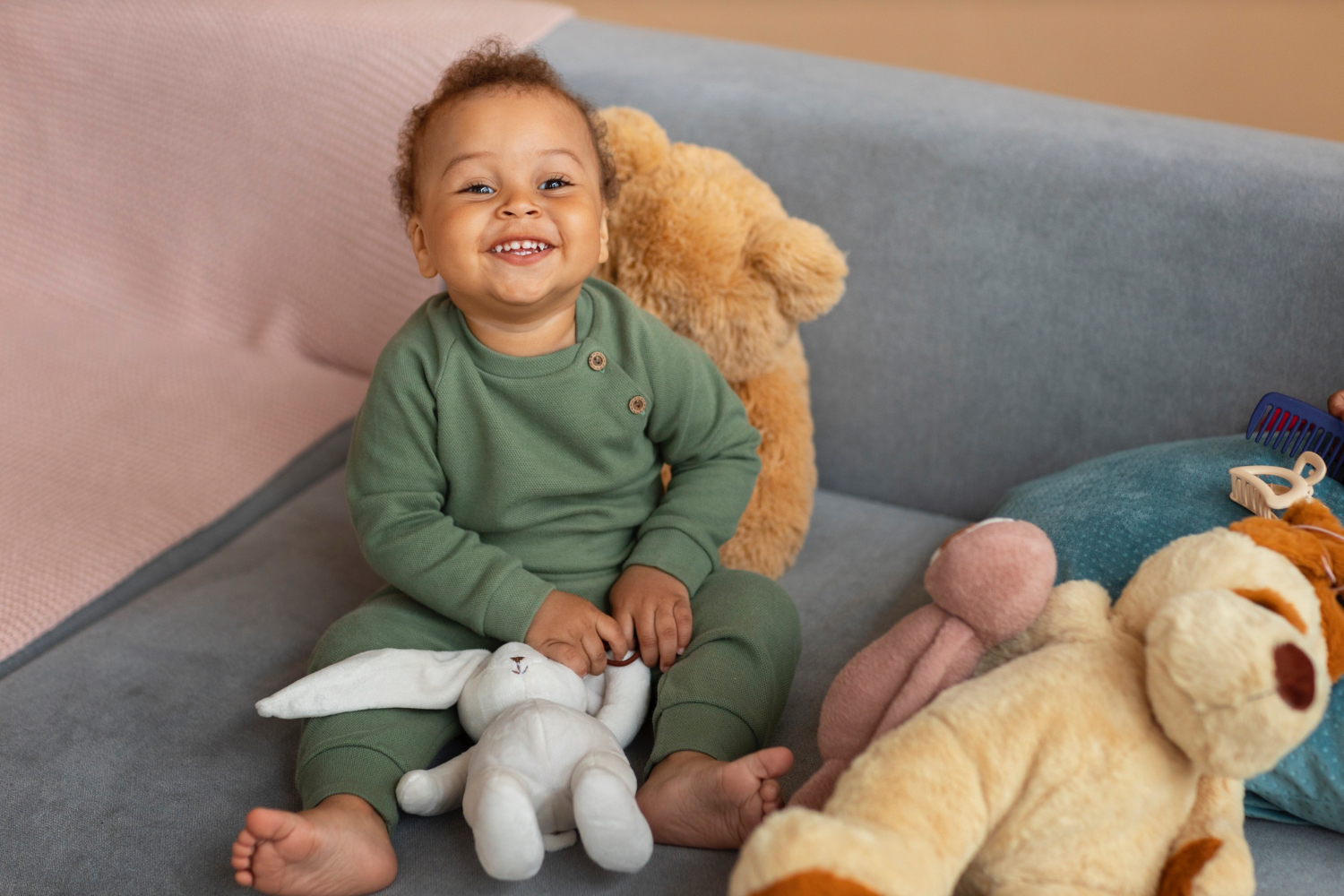 Toddler wearing dark green slitting on couch with multiple stuffed animals smiles at the camera.