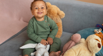 Toddler wearing dark green slitting on couch with multiple stuffed animals smiles at the camera.