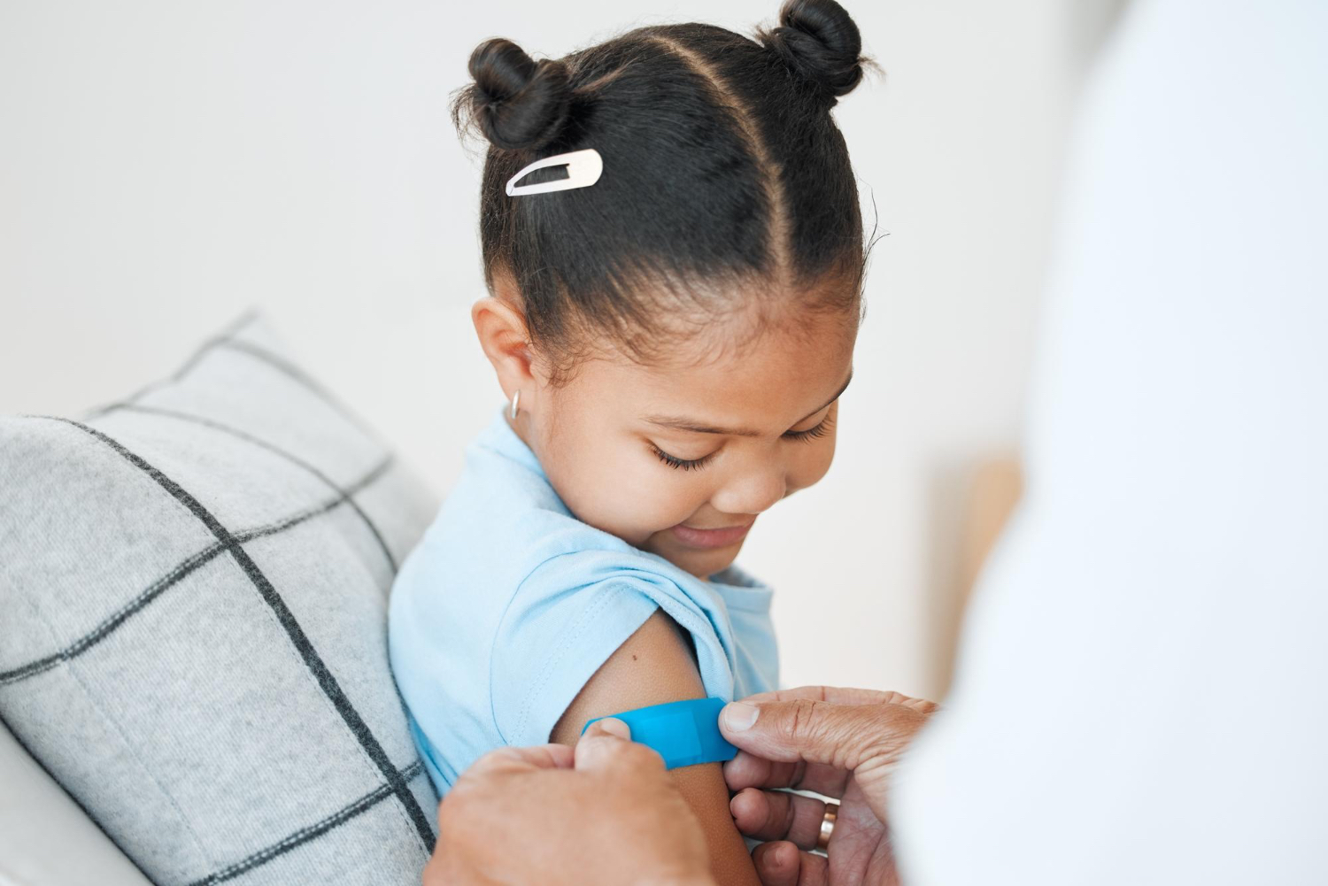 Young girl sitting and smiling gets a band aid put on her right arm by doctor figure, wearing blue t-shirt 