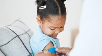 Young girl sitting and smiling gets a band aid put on her right arm by doctor figure, wearing blue t-shirt 