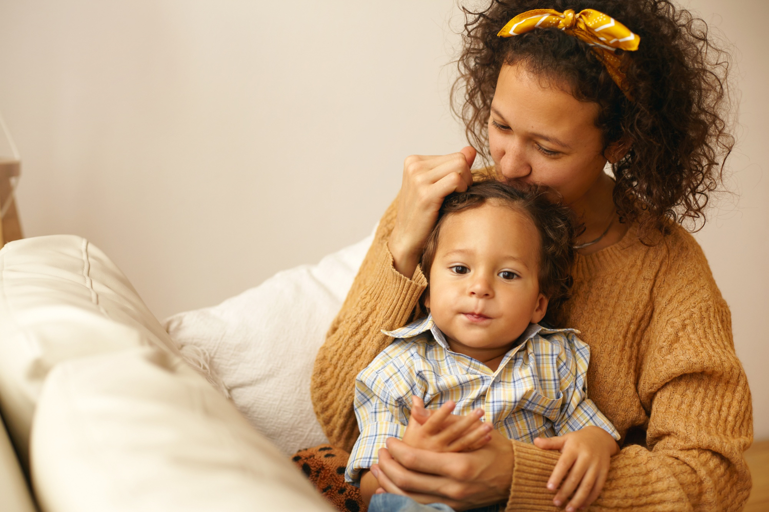 Adult female in cozy home setting holds toddler in her lap while kissing the top of his head. 