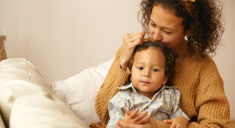 Adult female in cozy home setting holds toddler in her lap while kissing the top of his head. 