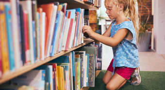 Little girl looks through books on big bookshelf in library setting.