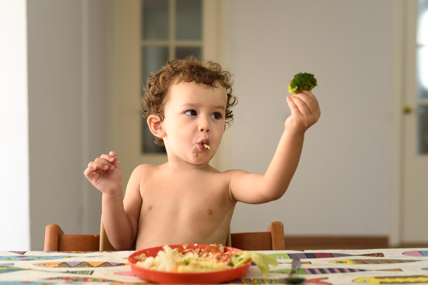 Little boy sitting at dinner table shirtless holding a piece of broccoli up in the air with a plate of food on the table. 