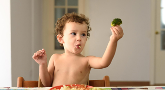 Little boy sitting at dinner table shirtless holding a piece of broccoli up in the air with a plate of food on the table. 