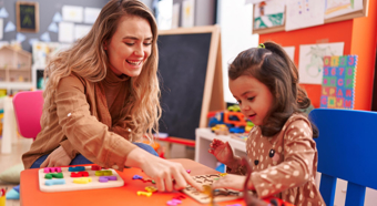Adult female sitting with toddler in a colorful classroom setting, helping the toddler with a puzzle. 