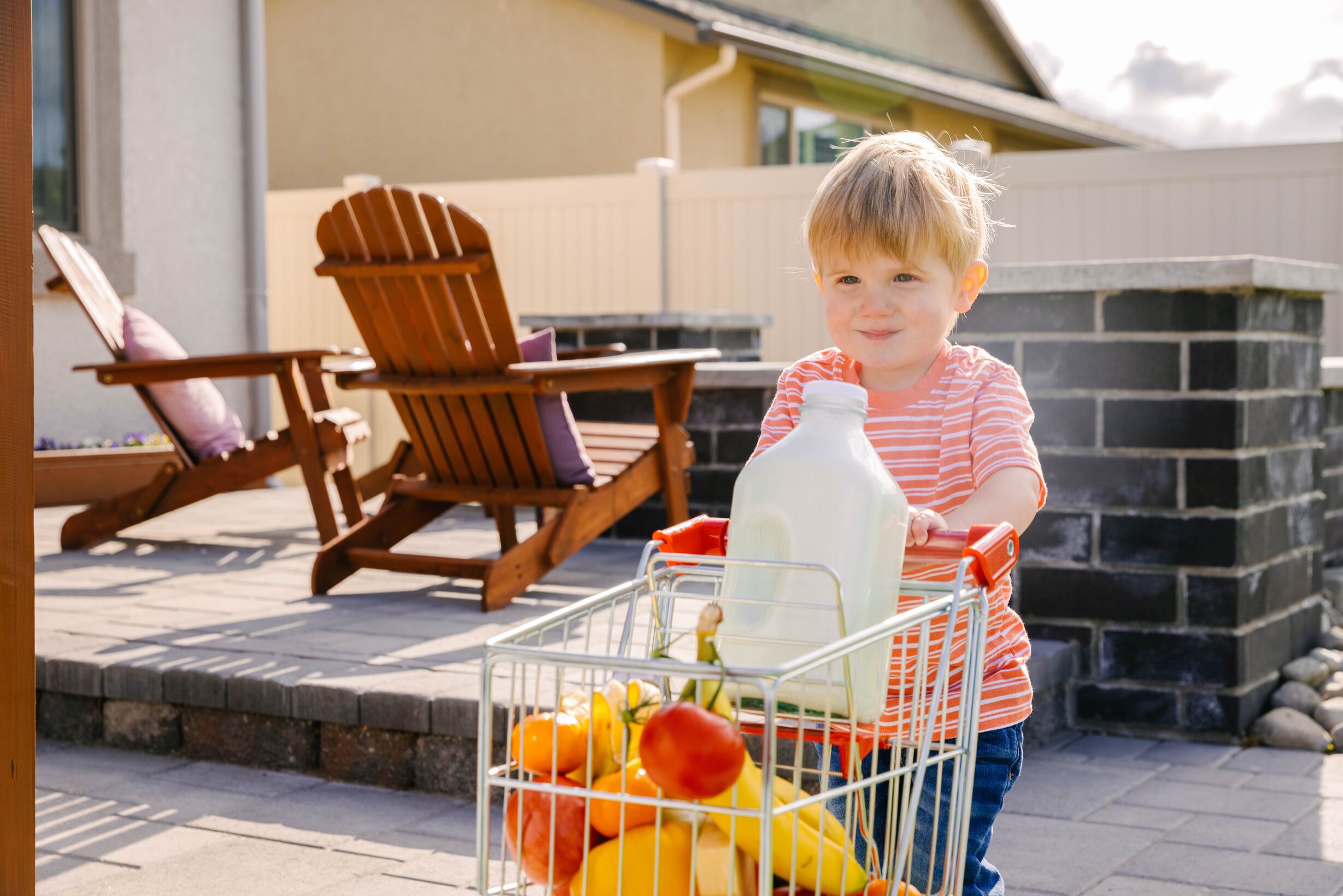 Young toddler boy wearing orange striped shirt, pushing a mini shopping cart with fruits and a big container of milk.