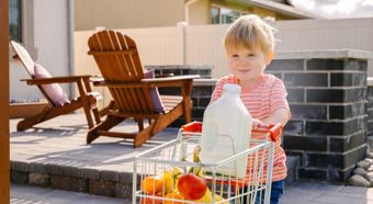 Young toddler boy wearing orange striped shirt, pushing a mini shopping cart with fruits and a big container of milk.