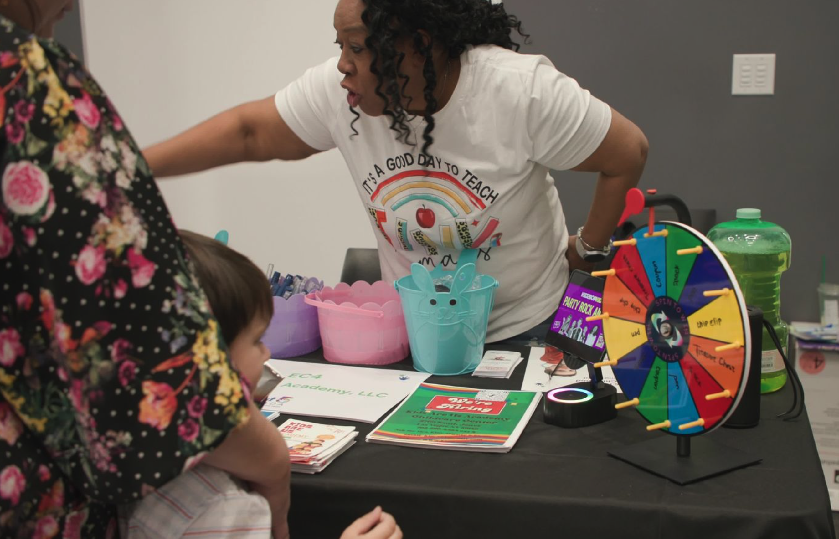 Toddler and adult watch lady at an event table demonstrate her booth and the spinning wheel on the table. 