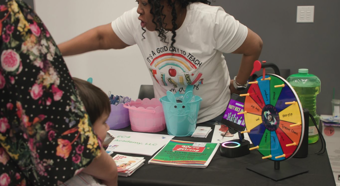 Toddler and adult watch lady at an event table demonstrate her booth and the spinning wheel on the table. 
